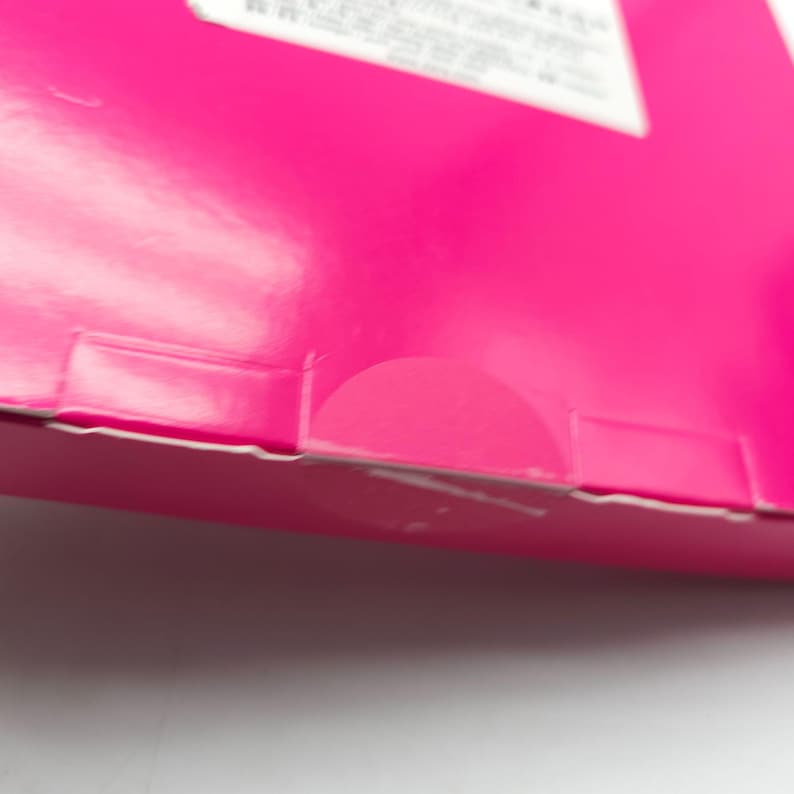 K&ouml;nnte beinhalten: Nahaufnahme einer leuchtend rosa Pappschachtel mit einem teilweise sichtbaren wei&szlig;en Etikett. Die Schachtel hat eine gefaltete Kante und einen halbkreisf&ouml;rmigen Ausschnitt. Die Oberfl&auml;che ist gl&auml;nzend und reflektiert Licht.
