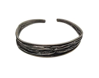Puede incluir: Un brazalete de plata con un diseño texturizado y en capas. El brazalete está abierto y tiene un borde liso y redondeado.