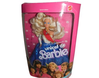 May include: A Special Edition UNICEF Barbie doll in its original pink box. The doll has blonde hair, blue eyes, and wears a white top, a red sash, and a blue skirt with white stars. The box features the UNICEF logo and images of children.