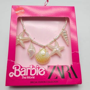 K&ouml;nnte beinhalten: Eine Barbie The Movie Special Edition Halskette mit Muschel-Anh&auml;ngern. Die Halskette hat eine Kette aus wei&szlig;en Perlen mit schillernden Seestern-, Jakobsmuschel- und Schneckenanh&auml;ngern. Die Halskette wird in einer rosa Schachtel mit den Logos von Mattel und Zara pr&auml;sentiert.