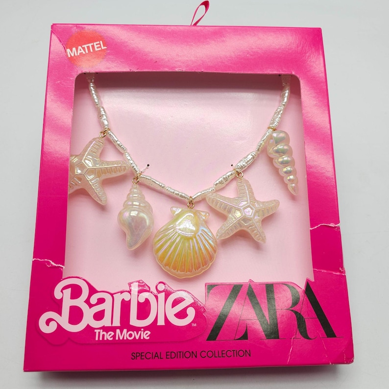 K&ouml;nnte beinhalten: Eine Barbie The Movie Special Edition Halskette mit Muschel-Anh&auml;ngern. Die Halskette hat eine Kette aus wei&szlig;en Perlen mit schillernden Seestern-, Jakobsmuschel- und Schneckenanh&auml;ngern. Die Halskette wird in einer rosa Schachtel mit den Logos von Mattel und Zara pr&auml;sentiert.