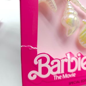 K&ouml;nnte beinhalten: Nahaufnahme einer rosa Schachtel mit dem Wort "Barbie" in wei&szlig;er Schrift. Die Schachtel hat einen eingerissenen Rand. Iridescent Muschelf&ouml;rmige Charms h&auml;ngen an einer Schnur. Der Text "The Movie" und "Special Edition" sind ebenfalls sichtbar.