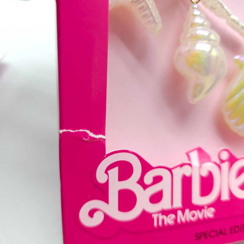 K&ouml;nnte beinhalten: Nahaufnahme einer rosa Schachtel mit dem Wort "Barbie" in wei&szlig;er Schrift. Die Schachtel hat einen eingerissenen Rand. Iridescent Muschelf&ouml;rmige Charms h&auml;ngen an einer Schnur. Der Text "The Movie" und "Special Edition" sind ebenfalls sichtbar.