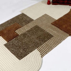May include: A rectangular area rug featuring a geometric composition of cream, dark brown, and tan sections. The design incorporates textured cream braided borders, a dark brown grid-patterned panel, and a central section with thin brown stripes.