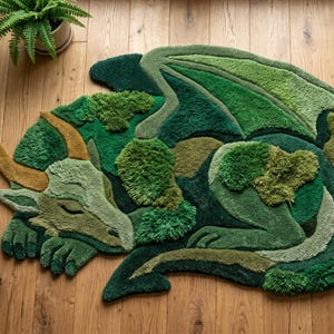 May include: A tufted rug shaped like a sleeping dragon, featuring various shades of green and mossy textures. The dragon has a curved golden horn, closed eyes, and a long tail, with a layered, plush surface that mimics a natural, forest-like appearance.