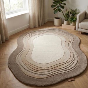 May include: An oval rug in shades of beige and brown, with a layered, topographic design. The rug's surface features concentric, wavy lines, creating a textured effect. It is placed on a wooden floor, adding warmth to the room.