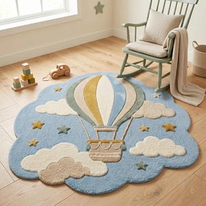 May include: A cloud-shaped area rug in light blue featuring a central hot air balloon in shades of cream, yellow, and sage green. The rug is decorated with tufted cream-colored clouds and small yellow and sage green stars scattered across the surface.