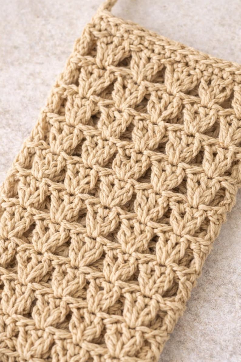 Pu&ograve; includere: Primo piano di un oggetto beige all'uncinetto con un motivo ripetuto a forma di foglia. Il pezzo fatto a mano ha una superficie strutturata e un anello nella parte superiore, che suggerisce che potrebbe essere una maniglia o un anello per appenderlo. Il colore neutro lo rende versatile.