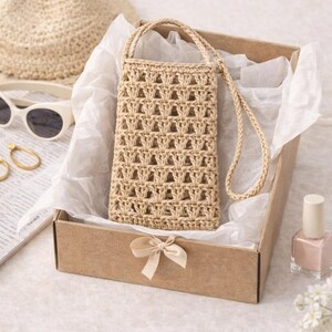 Pu&ograve; includere: Una borsa per telefono all'uncinetto beige con una lunga tracolla, presentata in una scatola marrone con un fiocco. Sono visibili anche un cappello di paglia, occhiali da sole e smalto per unghie, suggerendo un tema estivo.