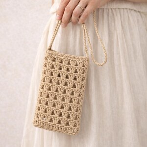 Pu&ograve; includere: Una borsa per telefono beige all'uncinetto con una lunga tracolla. La borsa ha una forma rettangolare con un design a trama aperta. La tracolla &egrave; realizzata con lo stesso materiale della borsa. La borsa &egrave; tenuta da una persona che indossa un vestito bianco.