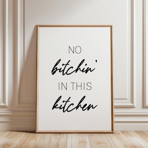 May include: A framed print with the words "NO bitchin' IN THIS kitchen" in black script. The artwork is in a wooden frame and is set against a white wall with moulding, on a wooden floor.