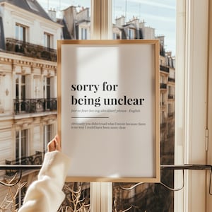 以下が含まれることがあります： 「sorry for being unclear」というテキストが太字のモダンなフォントで書かれた額入りのプリント。プリントには発音記号と説明文が含まれています。フレームは明るい木の色で、背景にはヨーロッパ風の建物が写っています。
