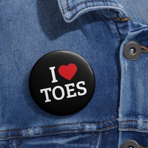 Puede incluir: Un botón negro con el texto "I ❤ TOES" en blanco, con un corazón rojo que reemplaza la palabra "love". El botón está sobre un fondo de mezclilla azul.
