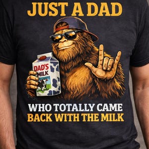Op de afbeelding: Zwart t-shirt met de tekst "JUST A DAD WHO TOTALLY CAME BACK WITH THE MILK". De afbeelding toont een Bigfoot-achtig wezen met een zonnebril en een baseballpet, die een pak "DAD'S MILK" vasthoudt en een handgebaar maakt.
