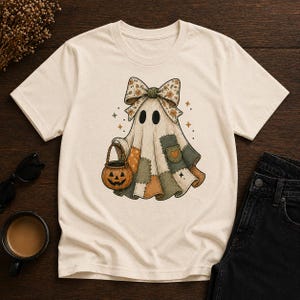 Könnte beinhalten: Cremefarbenes T-Shirt mit einem skurrilen Geisterdesign. Der Geist trägt einen Flickenteppich-Umhang, eine Blumen-Schleife und trägt einen Kürbis-Eimer. Das Design beinhaltet Sterne und ein Herz. Perfekt für Halloween oder die gruselige Jahreszeit.