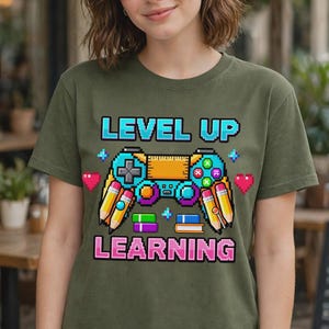 May include: An olive green t-shirt featuring a pixel art graphic of a video game controller combined with school supplies. The text reads LEVEL UP LEARNING in a pixelated font. The design includes pencils, books, and hearts in vibrant colors.