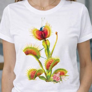 May include: White t-shirt featuring a colorful illustration of a Venus flytrap plant. The graphic shows the plant's distinctive red and green traps, with a detailed depiction of the plant's structure. The design is centered on the shirt.