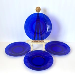 May include: Four cobalt blue glass plates with concentric ridged detailing. One plate is displayed on a small, ornate stand, with three additional plates arranged in front. The plates have a dark center ring.