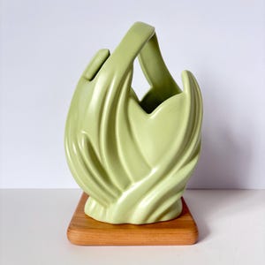 May include: A light green ceramic basket-style vase with a handle. The vase has a unique, flowing design and sits on a small, rectangular wooden base. The background is white.