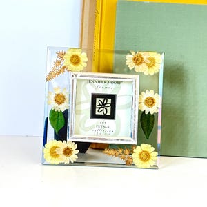 Op de afbeelding: Vierkante, heldere fotolijst van hars met ingebedde gele madeliefjes en groen. Een kleiner wit frame binnenin toont de tekst "JENNIFER MOORE frames the PETALS collection 6,35 x 6,35 cm". De lijst wordt weergegeven voor gele en groene boeken.