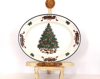 Puede incluir: Una bandeja de servicio ovalada de cerámica con una ilustración central de un árbol de Navidad con regalos debajo. El borde está decorado con cintas rojas, campanas doradas y hojas de acebo verdes. La bandeja descansa sobre un soporte de madera crema.