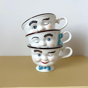 May include: Three stacked white ceramic mugs with painted faces, each with a different expression. The mugs have brown rims, handles, and the word "Baileys" on them. The bottom mug has a blue bow tie.