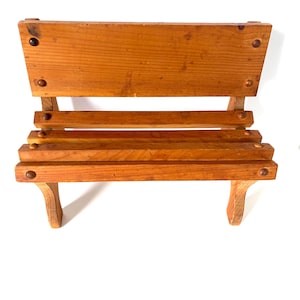 May include: A miniature wooden bench, featuring a backrest and two seat slats. Constructed from light brown wood, the bench has rounded legs and is held together with dark pegs. This decorative bench is designed for display purposes, with a classic design.