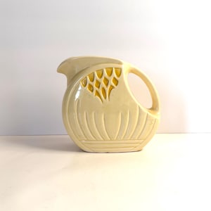 May include: Cream-colored ceramic pitcher with a rounded body and a handle. The pitcher features a decorative yellow design on the front and vertical grooves along the base. The spout is curved, and the handle is a simple loop.