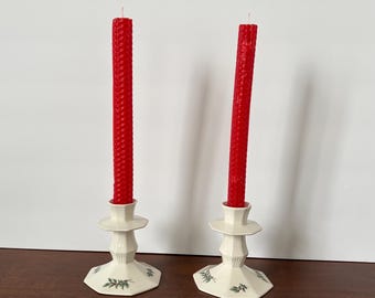 May include: Two off-white ceramic candle holders with an octagonal base and a faceted stem. Each holder features a small green and red holly leaf pattern on the base and holds a tall, textured, bright red beeswax taper candle.