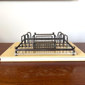 Op de afbeelding: Een vintage metalen brievenhouder met een rechthoekig ontwerp, met meerdere compartimenten en een decoratief frame. De brievenhouder is geplaatst op een lichtgeel boek, waardoor een klassiek bureauaccessoire ontstaat.