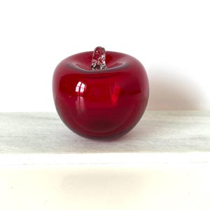 Puede incluir: Una manzana decorativa de cristal rojo brillante con un tallo transparente y texturizado. La pieza decorativa es de un tono rojo vibrante y se encuentra sobre una superficie blanca. La manzana tiene una forma sólida y redondeada y refleja la luz.