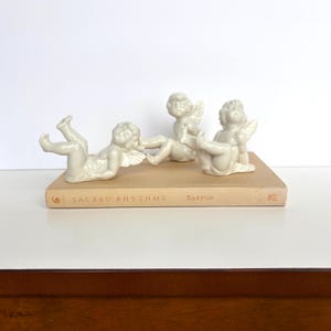 Könnte beinhalten: Drei cremefarbene Keramik-Puttenfiguren in verschiedenen Posen liegen auf einem beigefarbenen Buch. Auf dem Buchdeckel steht "SACRED RHYTHMS" und "BARTON". Die Putten haben detaillierte Flügel.