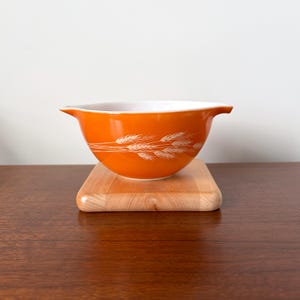 May include: An orange mixing bowl with a white interior and a wheat stalk design. The bowl is sitting on a small wooden cutting board. The bowl has a handle on each side.