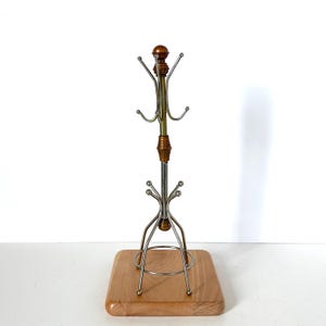 Può includere: Un supporto per tazze con base quadrata in legno chiaro e asta centrale in metallo. Il supporto presenta due livelli di ganci metallici argentati curvi con estremità arrotondate, arricchiti da distanziatori cilindrici color rame a coste.