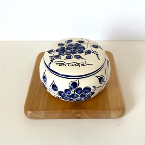 May include: A round ceramic lidded jar with a cream-colored base and hand-painted dark blue floral patterns. The lid features the word Portugal written in black script. The jar has a smooth, glossy finish and a rounded, squat silhouette.