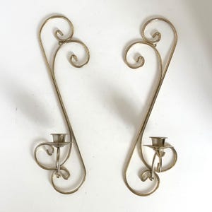 May include: A pair of wall-mounted candle holders crafted from polished silver-toned metal. Each piece features a long, slender, curved frame with decorative scrollwork at the top and a circular cup designed to hold a single taper candle.