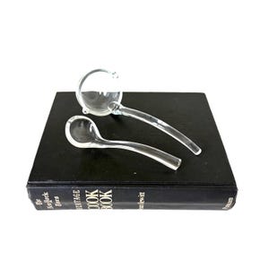 May include: Two clear glass ladles, one with a round bowl and the other with a spoon-shaped bowl, rest on a black book. The book is titled "The New York Times Heritage Cook Book" and is a cookbook.