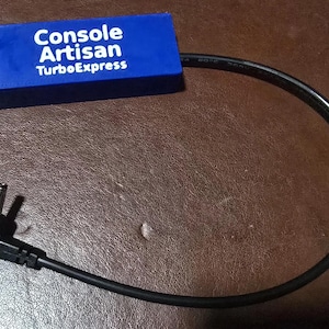May include: A blue rectangular power adapter block with the white text Console Artisan TurboExpress printed on the top surface, featuring two black screws on the left side and a black power cable extending to an L-shaped barrel connector plug.