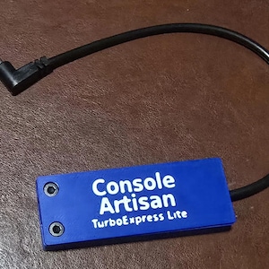 May include: A rectangular blue electronic component with two visible screws on the left side. The text Console Artisan TurboExpress Lite is printed in white on the surface. A black power cable with an L-shaped connector extends from the unit.