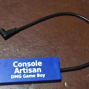 May include: A rectangular blue electronic accessory with white text that reads Console Artisan DMG Game Boy. The device features two black circular screw heads on the left side and is connected to a black power cable with an L-shaped plug.