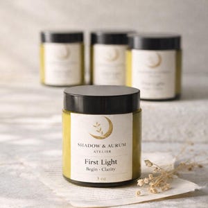 May include: Four glass jars with black lids, filled with a yellow substance. The front jar has a label that reads "SHADOW & AURUM ATELIER First Light Begin • Clarity 3 oz."
