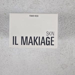 May include: A white rectangular box with the text "POWER REDO" at the top and "IL MAKIAGE SKIN" in bold black letters. The box is centered on a textured, light gray surface.