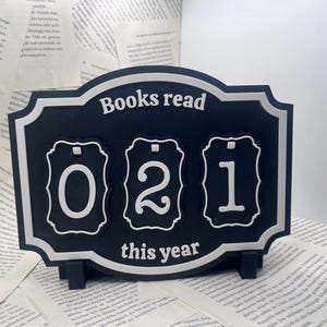 Könnte beinhalten: Ein schwarz-weißes rechteckiges Schild mit dekorativen Rändern, das oben den Text "Books read" und unten "this year" trägt. Drei kleinere, passende rechteckige Plaketten mit den Ziffern 0, 2 und 1 sind daran befestigt.