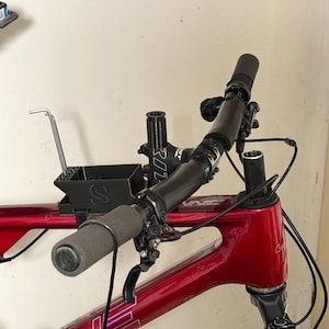 Può includere: Primo piano di un telaio di bicicletta rosso con manubrio e componenti neri. Il manubrio ha impugnature grigie. Un contenitore nero è attaccato al telaio. La bicicletta è contro un muro neutro.