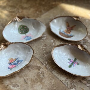 May include: Four decorative seashell dishes with gold edging. Each shell has a white interior with a different Easter-themed design: a tree, a bunny in a basket, a chick in a teacup, and a bow.