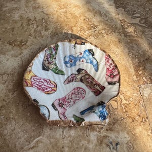Könnte beinhalten: Eine dekorative, muschelförmige Schale mit weißem Innenraum und bunten Cowboy-Stiefel-Designs. Die Stiefel sind in verschiedenen Farben gehalten, darunter Rosa, Blau und Grün, mit goldenem Rand.