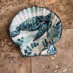 Puede incluir: Un plato decorativo con forma de concha marina con un diseño de tortuga marina turquesa. La tortuga está pintada en tonos turquesa y azul, rodeada de plantas acuáticas. El plato tiene un fondo blanco con un borde brillante turquesa.