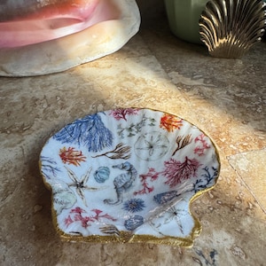 May include: A seashell-shaped decorative dish with a white interior featuring a colorful sea life design. The design includes seahorses, starfish, and coral in shades of blue, red, and orange. The dish has a gold-painted rim.