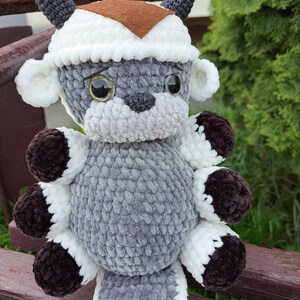 May include: A handmade plush toy in the shape of a bull, crafted from soft, textured yarn. The toy features a gray body, white accents, and brown details on the hooves and horns. It wears a white and brown hat, and has large, expressive eyes.