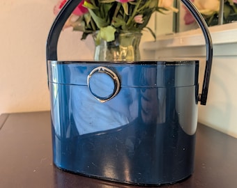May include: A vintage navy blue handbag with a curved handle. The oval-shaped bag has a shiny finish and a decorative clasp. The bag is sitting on a dark wooden surface. A bouquet of pink and purple flowers is in the background.